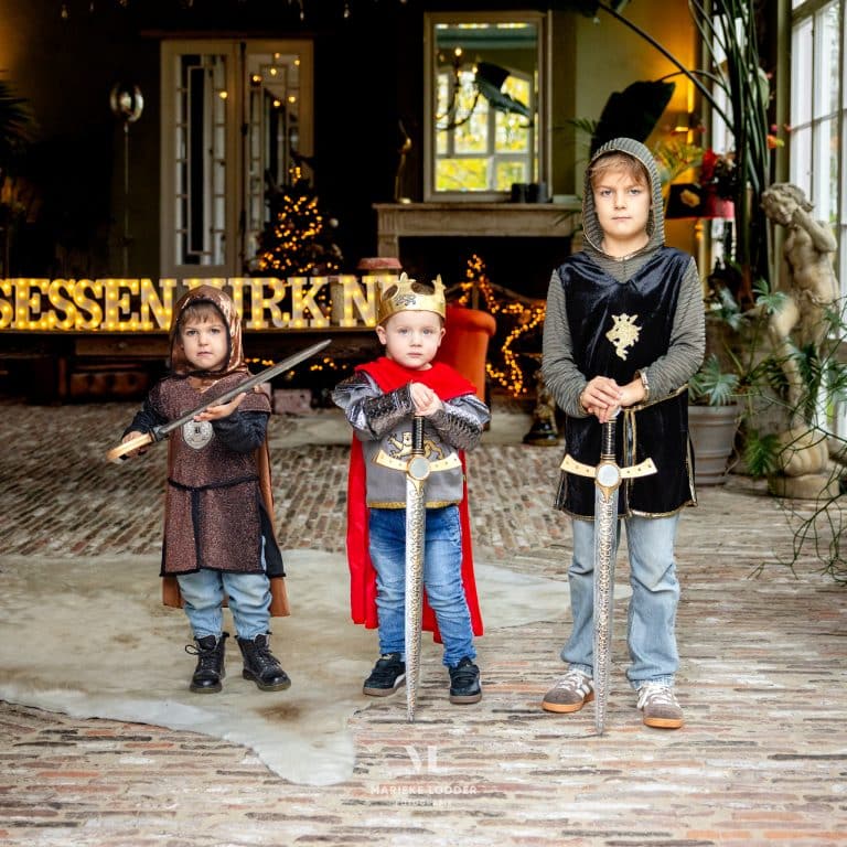 Jongen draagt het rode en bruine Arthur King ridder verkleedpak met kroon, rode cape en zwaard naast twee andere ridders