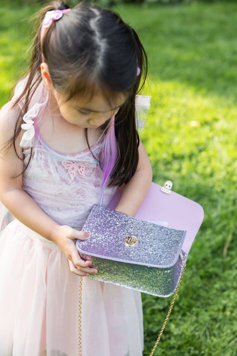 Meisje draagt een zilverkleurig glittertasje met paarse glitters, gouden ketting en sluiting in de tuin.