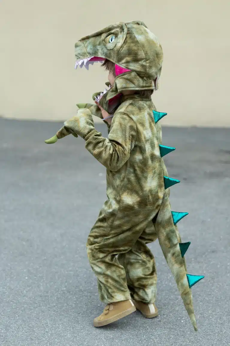 Kind in het Great Pretenders dino pak met dinosauruskap, klauwhanden, staart en turkooizen stekels.