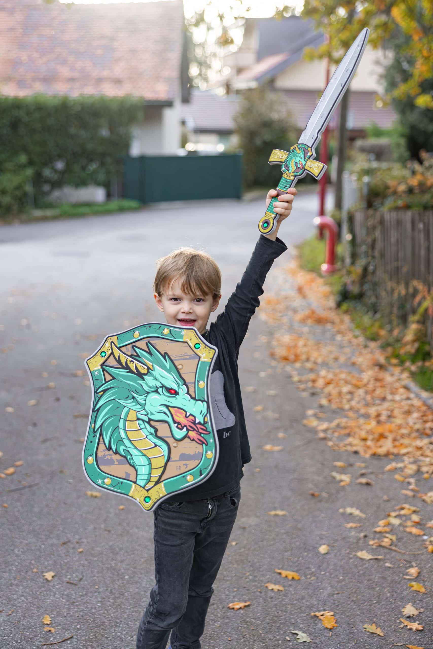 Jongen houdt het ridder-draken zwaard omhoog en draagt een ridders schild met een groene draak.