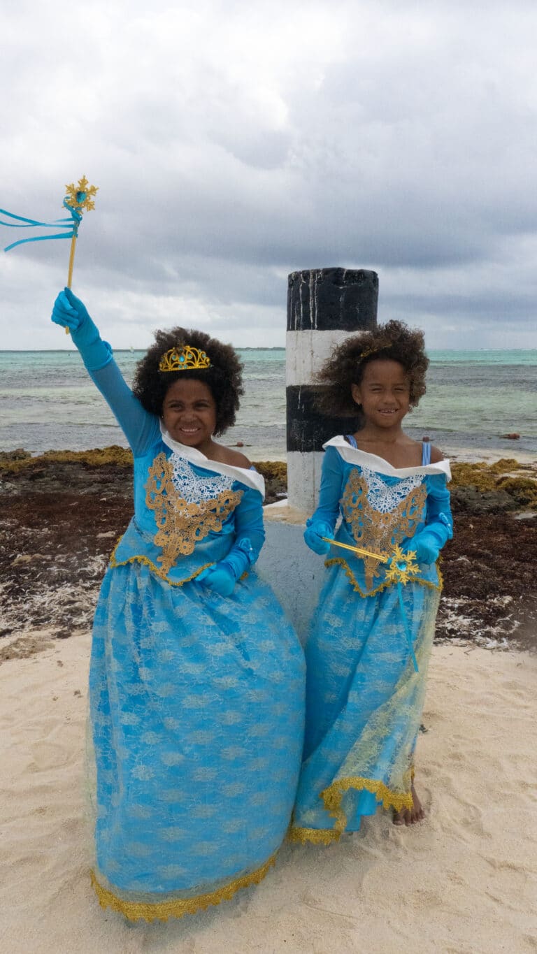 Twee meisjes dragen een blauwe assepoester jurk met gouden versieringen; één houdt een toverstaf vast op het strand.