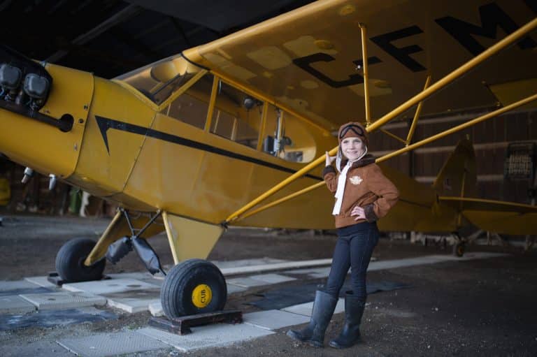 In een hangar poseert een kind in een bruin piloten pak kind naast een groot geel vliegtuig, met handen in de zij.