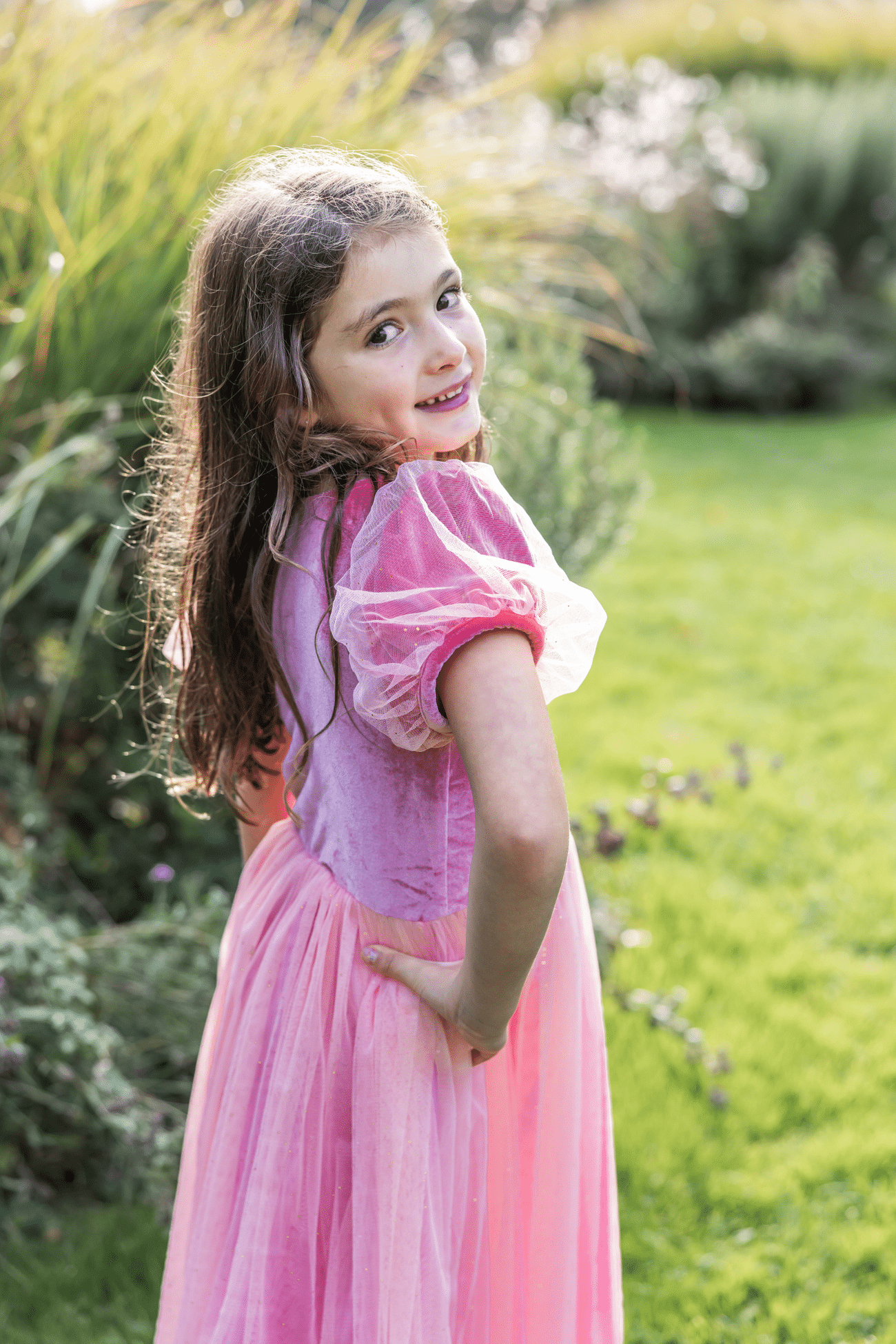 Meisje draagt een roze prinsessenjurk met tule rok en doorschijnende pofmouwen, zijwaarts kijkend in de tuin
