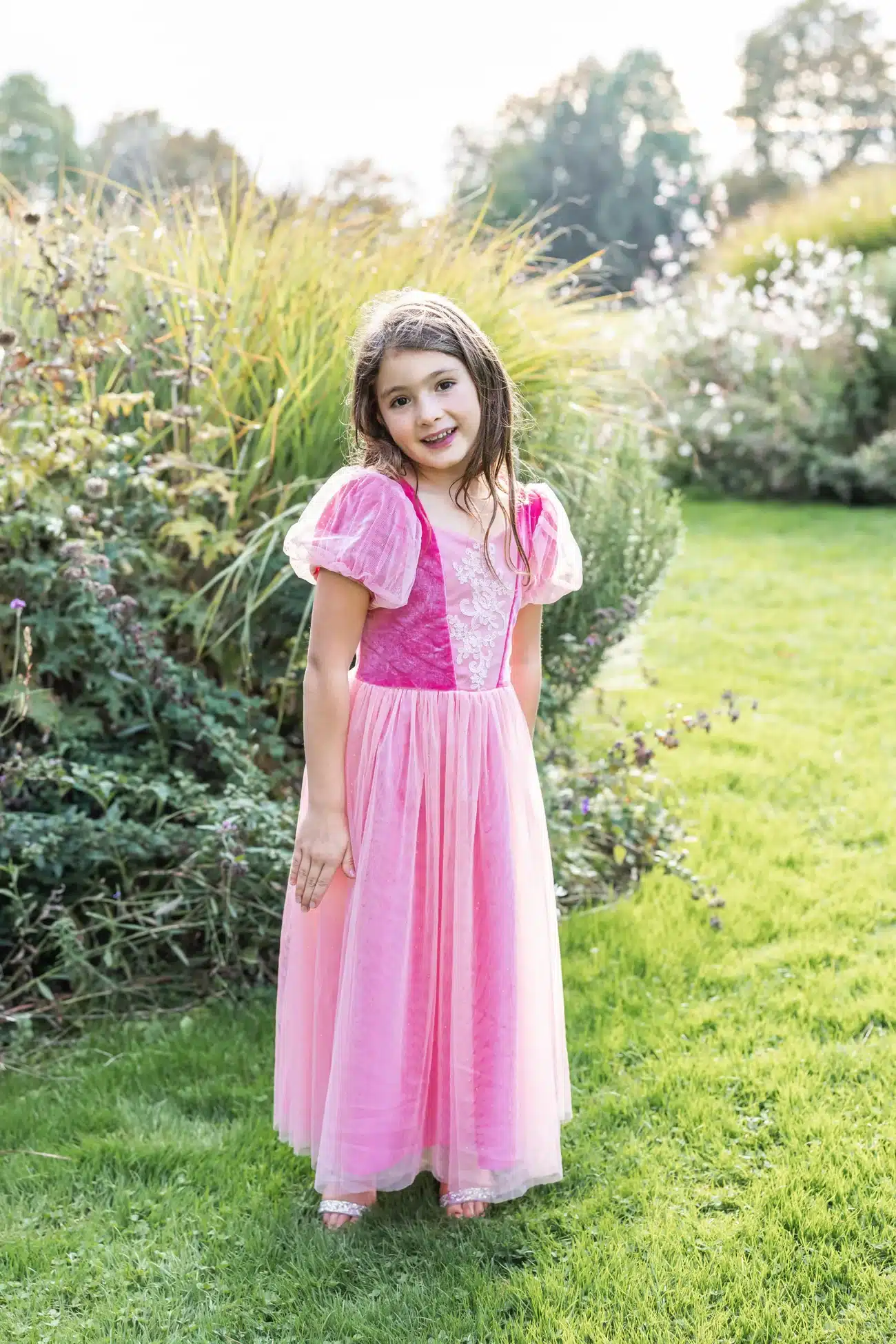 Meisje draagt een roze prinsessenjurk met doorschijnende pofmouwen en een lange tule rok in de tuin