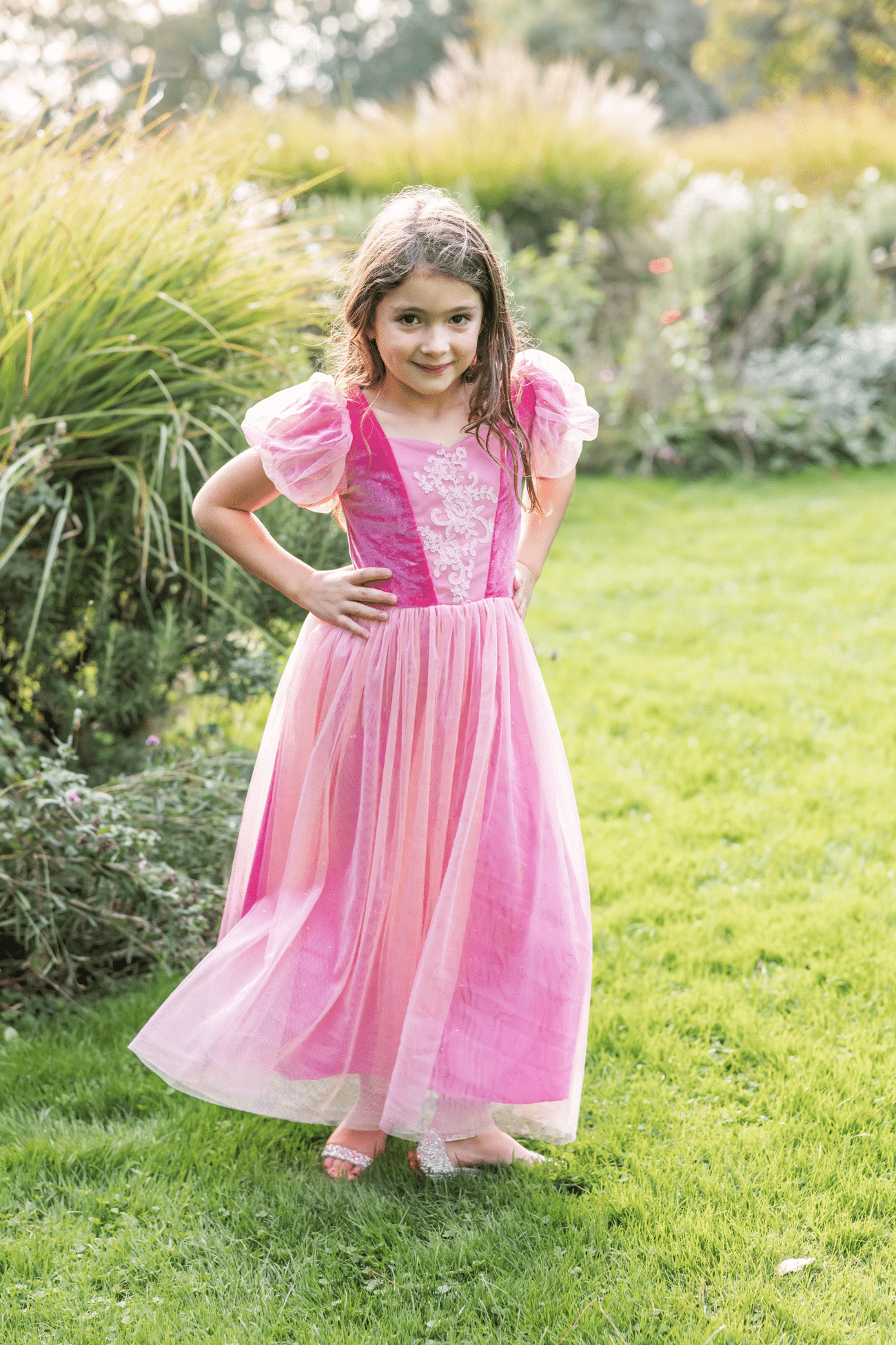 Van voren toont een meisje haar roze prinsessenjurk met geborduurd lijfje en handen in de zij, buiten in de tuin