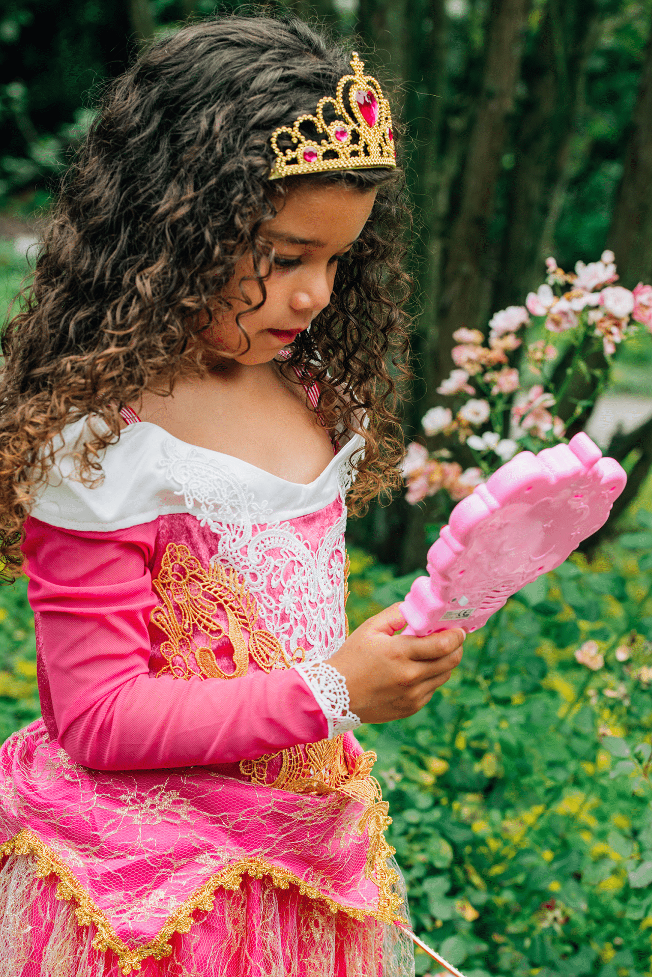 Meisje houdt een blauwe prinsessenspiegel met handgreep vast en bekijkt die in haar roze prinsessenjurk in de tuin.