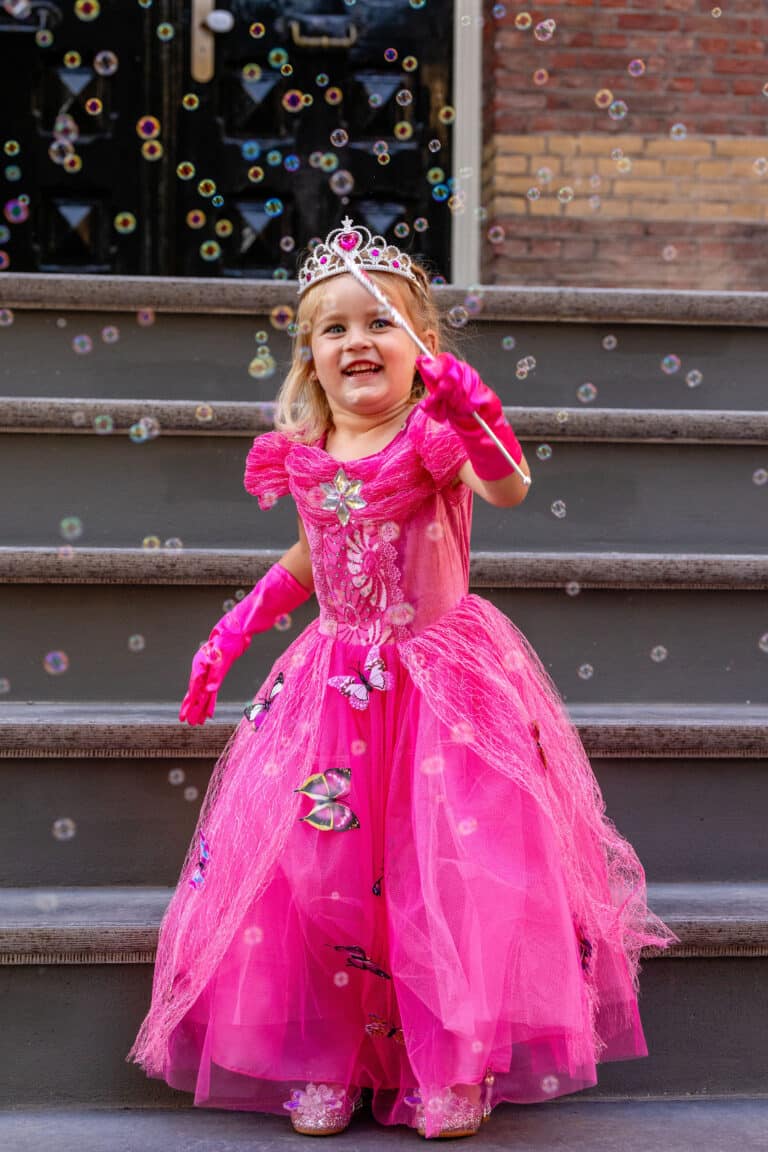 Meisje draagt een fuchsia jurk van Doornroosje met tiara, roze handschoenen, toverstaf en wijde tule rok op een trap.