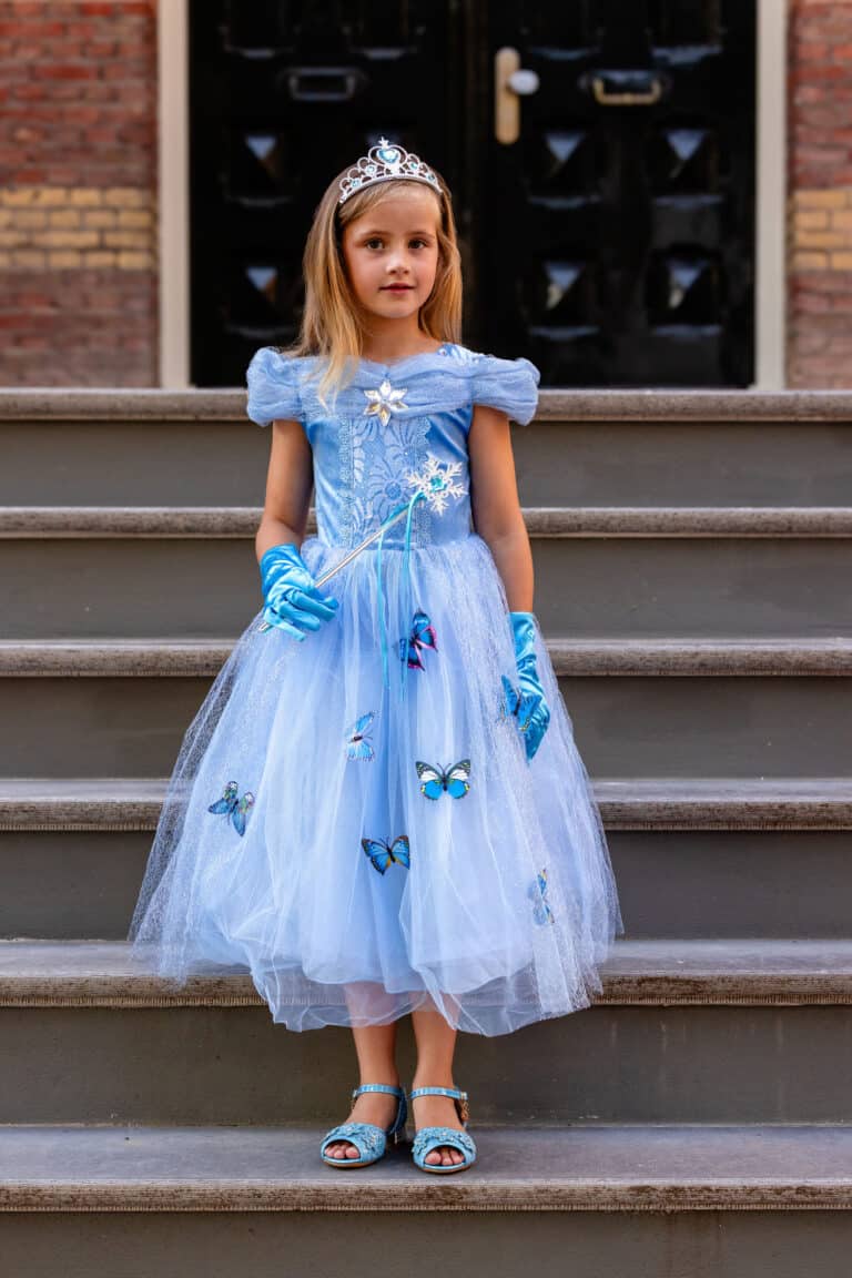 Meisje draagt een doornroosje blauwe jurk met tiara, handschoenen, toverstaf en vlinderdecoratie op een trap.