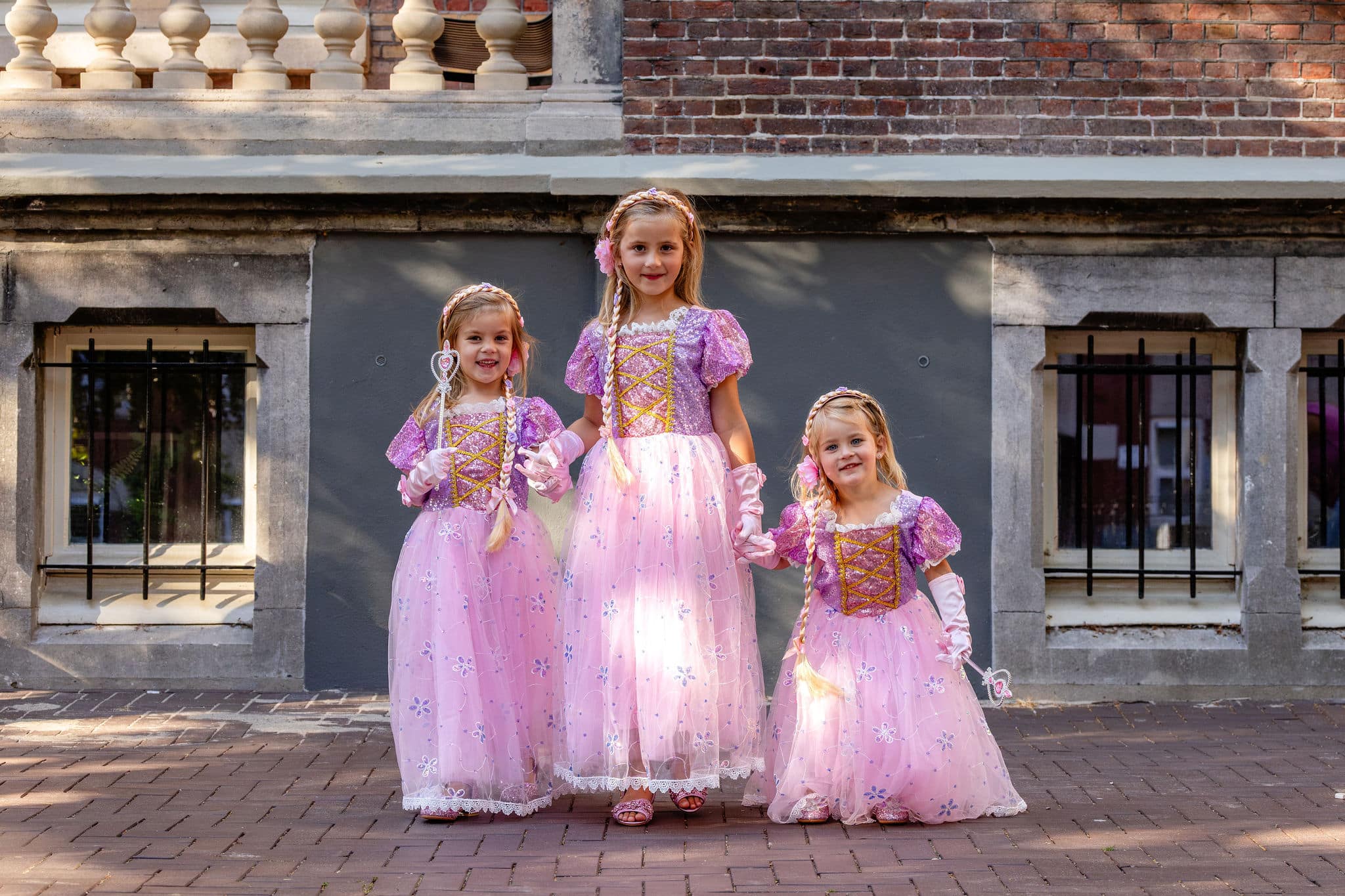 Drie meisjes dragen roze Rapunzel verkleedjurken met paarse glittertops, lange tule rokken en handschoenen voor een gebouw.